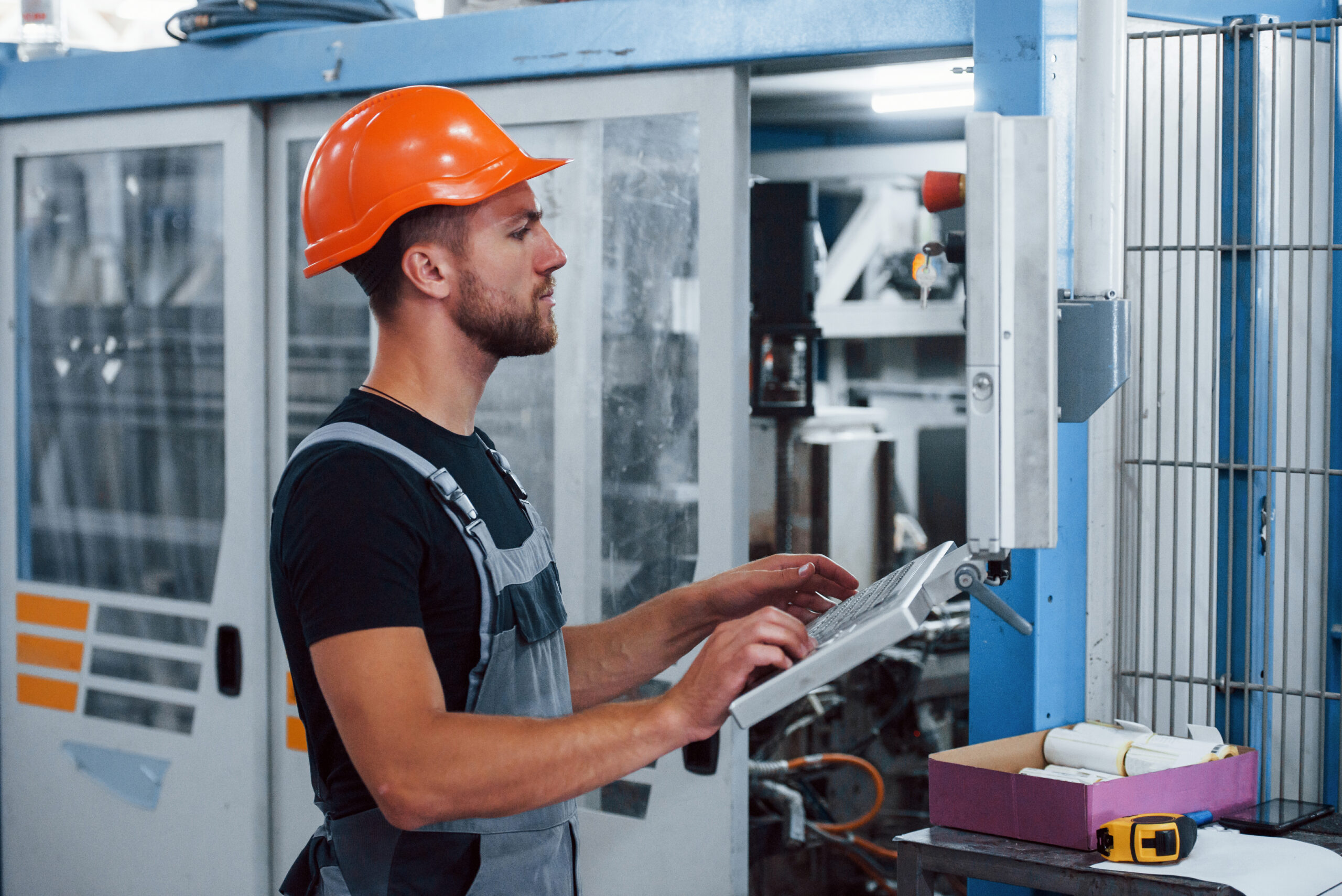 Using machine to program operations. Industrial worker indoors in factory. Young technician with orange hard hat